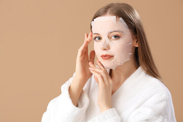 Young woman in bathrobe with facial sheet mask on beige background, closeup