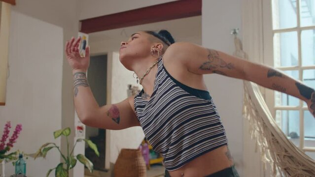 Young tattooed girl with piercing and undercut hairstyle dancing in sunlit living room with hammock during day at home