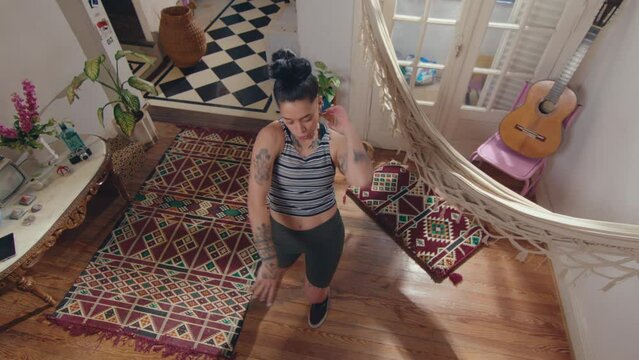 Young cool girl with tattoos and undercut hairstyle performing freestyle dance moves with inspiration, practicing new choreo in creatively designed living room with hammock. High angle view