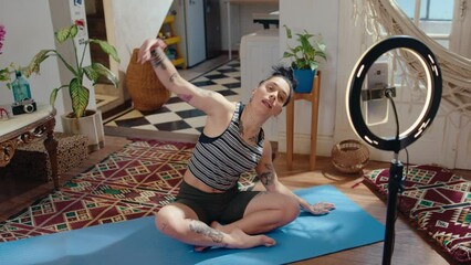 Young girl communicating with viewers, doing side bends and butterfly stretch in front of phone with ring light while filming yoga practice for fitness vlog at home
