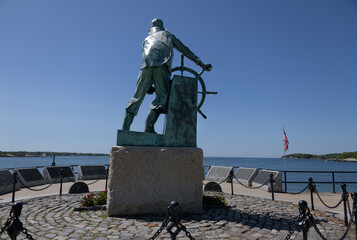 Bronze statue of a fisherman in oilskins standing braced at the wheel on the sloping deck of his...