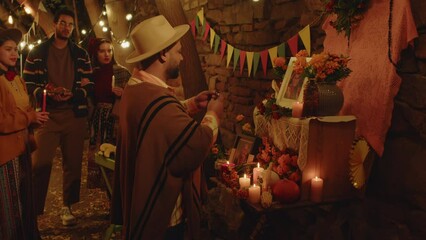 Medium shot of Hispanic man standing by Day of the Dead altar with praying hands and speaking to departed family member, leaving his rosary beads near framed picture of old happy woman