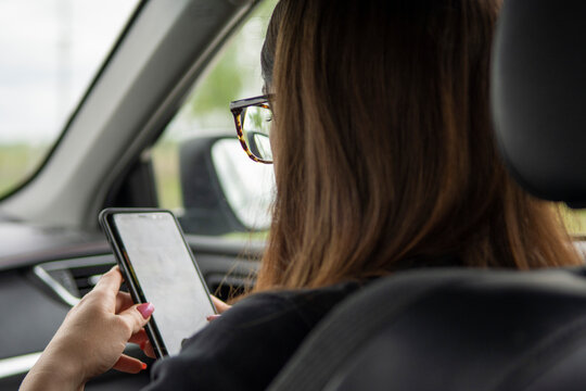 Brunette with long hair and glasses looks at phone in passenger seat of car