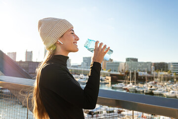 Happy sporty woman taking a break from running drinking water outdoors during winter cold weather...