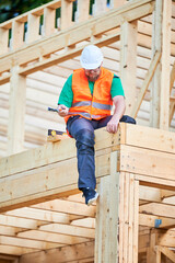 Carpenter constructing two-story wooden frame house. Bearded man hammering nails into structure while wearing protective helmet and construction vest. Concept of modern ecological construction.