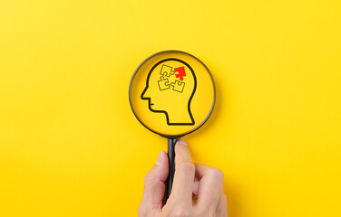 Hand holding magnifying glass focusing on human brain jigsaw puzzle on yellow background. human...