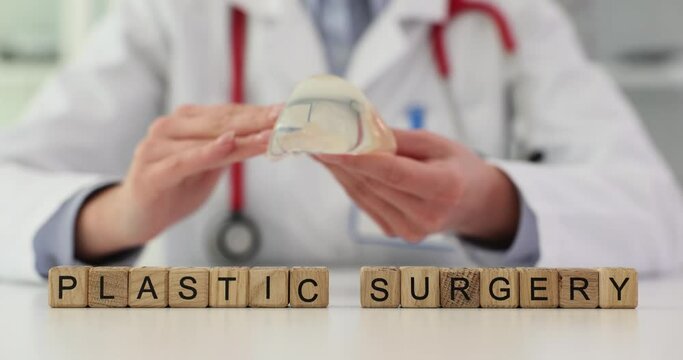 Doctor mammologist holding breast implant near word plastic surgery on wooden cubes 4k movie slow motion