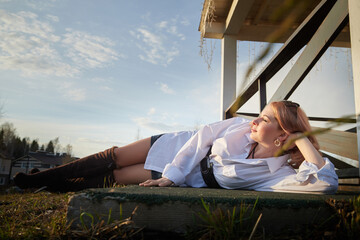 Beautiful girl with red hair in white shirt in open wooden pavillion in village or small town. Young slender woman and sky with clouds on background on an autumn, spring or summer day