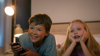 Smiling Caucasian children happy family kids boy girl brother sister siblings watching TV together watch cartoon show program using remote control enjoy television lying on bed in evening communicate