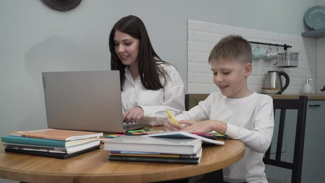 Medium shot happy mom is working on a laptop while sitting at the kitchen table, her son is doing homework next to her and helps mom figure out the laptop in parallel