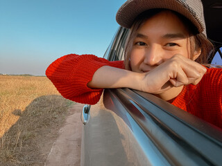 Asian Thai woman wear cap taking selfie, holding phone while sitting in the car as passenger, arm putting on car window, smiling happy having good time, traveling on vocation holidays. 