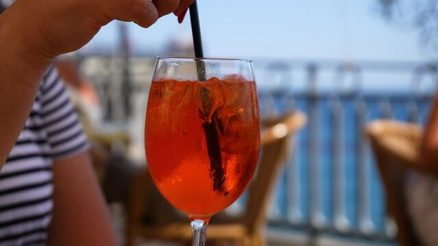 Closeup cropped shot of unrecognizable woman stirring aperol spritz cocktail with straw and drinking outdoors cafe on summer day on background of sea. Delicious alcoholic drink with citrus fruits.