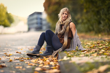 A blonde woman in knitted clothing sits on the side of the road, leans back and smiles into the...