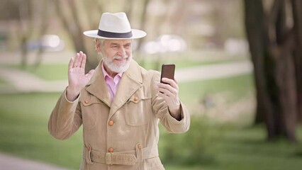 Front view of charismatic trendy pensioner in casual outfit waving hand and smiling during video chat by cell phone at public space. Caucasian old man learning using new mobile device for online call. - Powered by Adobe