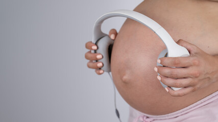 A pregnant woman holds headphones against her bare tummy. The expectant mother lets her child...
