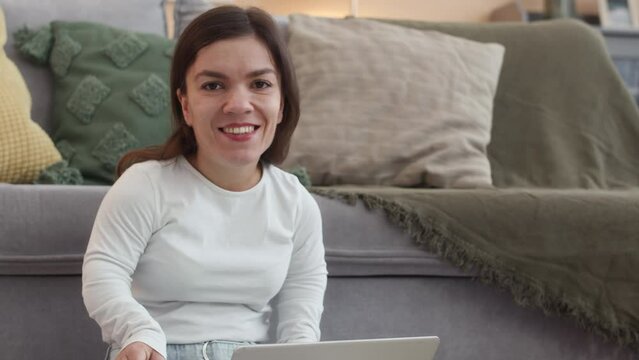 Slowmo portrait of young positive Caucasian woman with short stature sitting on floor with laptop on her thighs and smiling at camera while working from home