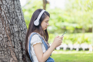 Teenage girl sitting against a tree trunk listening to music with headphones at park, Young Asian teen woman enjoying music