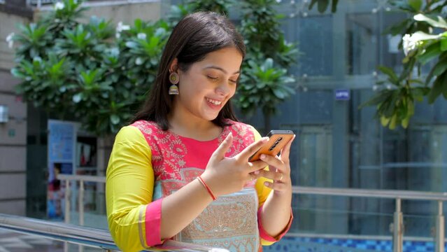 Young Indian woman using phone in a park - smiling  laughing  digital communication  overuse of phones. Brown girl texting a friend - gossiping  sharing jokes  text exchange  mobile chatting  smart...