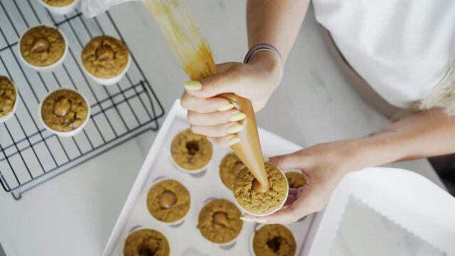 unrecognizable woman confectioner fills fresh muffin with caramel from plastic pastry bag making delicious natural dessert in kitchen at home close-up view flat-lay top with moving camera