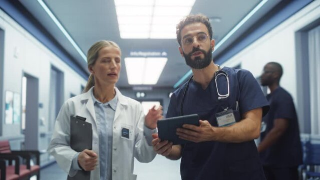 Portrait of Female Doctor and Male Nurse Walking in Hospital Corridor and Talking while Using a Digital Tablet. Medical Specialists Sharing Notes and Instructions, Planning a Surgery