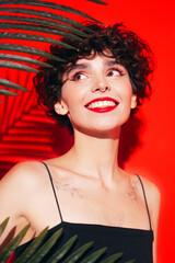 Fashion beauty portrait of young smiling brunette woman with evening stylish  makeup and perfect clean skin. Sexy model with curly short hair posing in studio. With red bright lips. Tropical palm leaf