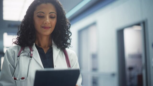 Portrait of a Female Doctor Wearing White Coat Working on Tablet Computer in Hospital Corridor. Medical Health Care Professional or Head Nurse Looks at the Camera and Gently Smiles. Slow Motion