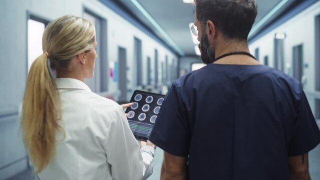 Backview Tracking Shot of Two Doctors Walking in Hospital Corridor, Using a Digital Tablet. Surgeon and Nurse Checking Brain MRI Images Before Medical Tumor Removal Intervention