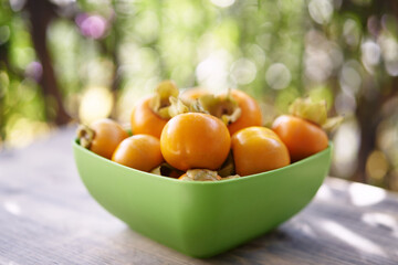 Full green bowl of ripe persimmons stands on a wooden table on the balcony