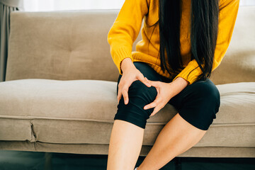 A young woman on sofa holding painful knee suffering chronic tendon arthritis. Health Care and...