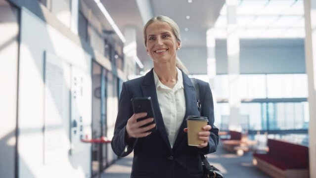 Portrait of a Caucasian Woman Using Her Smartphone, Walking in an Empty Office Hall and Smiling. A Businesswoman in Smart Casual Clothes Entering her Workplace in the Morning Holding a Coffee