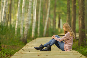 Sad young adult woman sitting alone on wooden trail at birch tree forest in autumn day. Thinking...
