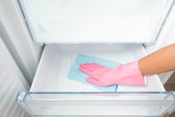 Young adult woman hand in pink rubber protective glove wiping inside plastic box of white freezer with dry blue rag. Closeup. Front view.