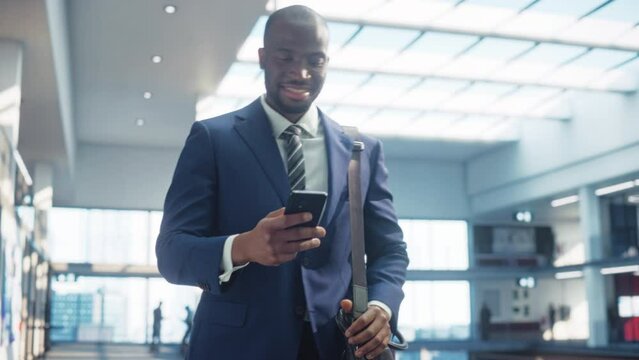 Portrait of Young Black Man in a Perfect Tailored Suit Walks in Corporate Office Hallway, Using his Smartphone and Smiling. Successful Businessman Communicating with Colleagues Over Text Messages