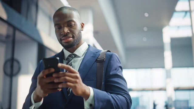 Portrait of Successful African-American Businessman Standing In Office Building Using Smartphone. Black Entrepreneur uses Mobile Phone e-Commerce App for Stock Market Investment Campaign. Slow Motion