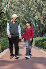 Naklejka premium Happy Indian grandparents holding playful grandson's hands and walking on footpath while spending weekend in park