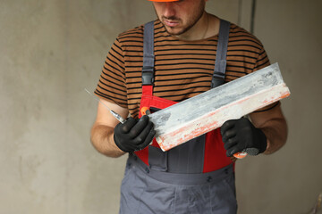 Worker in overalls and safety hard hat holds and prepares two spatulas for plastering concrete wall...