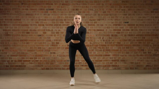 Fitness trainer performing Squat toe tap exercise. The girl is dressed in black sportswear and clearly demonstrates the physical exercise. She is against a brick wall in loft style. RED RAPTOR 8K RAW