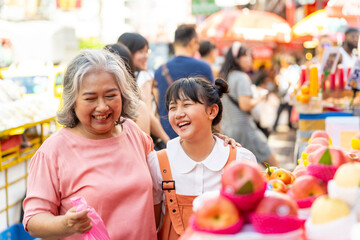 Happy Asian family grandmother and grandchild girl choosing and buying fresh fruit together at street market. Senior woman and little girl enjoy outdoor lifestyle travel in the city on summer vacation - obrazy, fototapety, plakaty