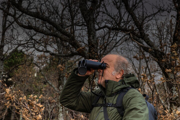 adult man with white hair and beard in the forest in winter, spotting birds with the binoculars he...