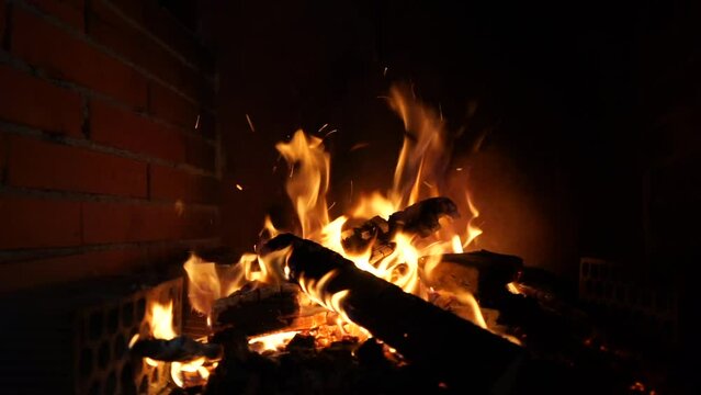 Beautiful video of the movement of fire flames and sparks at night against the background of bricks inside the fireplace. The magical movement of tongues of fire and flying sparks on a dark backgroun