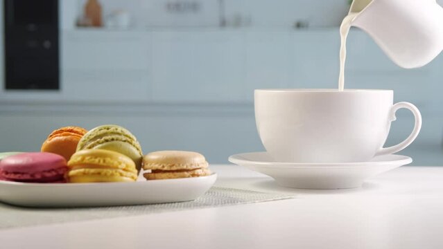 Latte. Woman pours milk from a milk jug into a coffee cup. Breakfast table in bright kitchen. Coffee with milk and colorful macarons. Sugar consumption and coffee consumption
