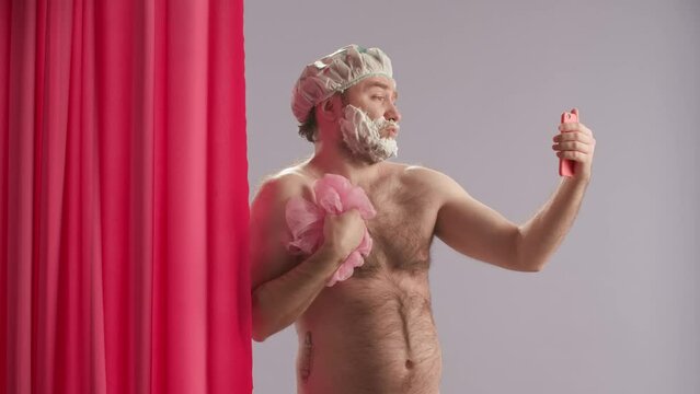 A man and with shaving foam on his face is taking a shower and using a smartphone. Man taking selfies, shooting video, streaming, blogging near pink shower curtain in studio on blue background.