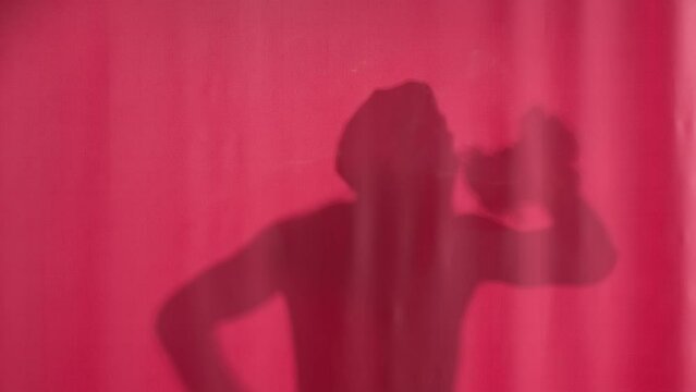 Silhouette of a man washing behind a pink curtain. Man in waterproof cap washing, dancing and singing, using loofah as a microphone. Body washing.