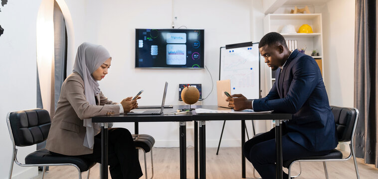 Beautiful Muslim woman talking on the phone and African man using smart phone while sitting at work place. Businessman sitting in modern office.
