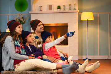 Happy parents with kids in warm winter wear watching tv or television at home during holidays - concept of weekend entertainment, family bonding and relationship. - obrazy, fototapety, plakaty