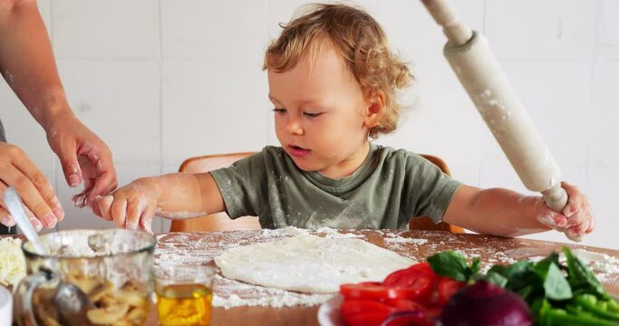 Joyful baby boy rolls out pizza dough with rolling pin, assisted by his mom, who scatters pinch of flour on flatbread. His enthusiasm grows as he takes matters into his own hands, pouring flour