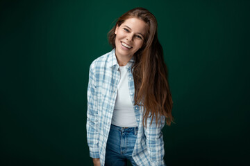 A young woman with brown hair wears a checkered blue shirt and stands on a green background with copy space