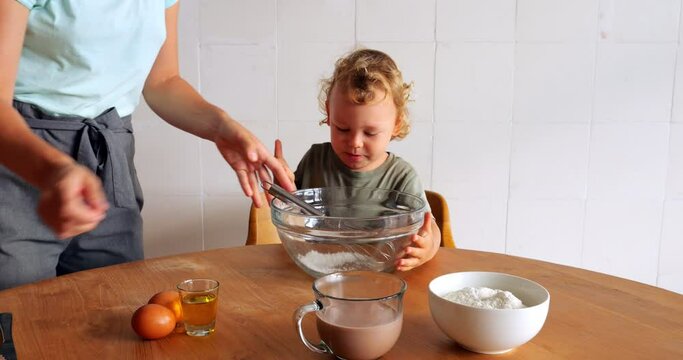 Impatient toddler eagerly stirs sugar in empty bowl with whisk, ready to start cooking pancakes, all captured in playful and adorable scene. Slow motion shot of young kitchen helper