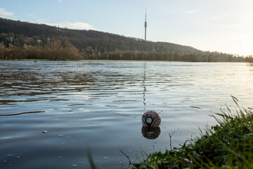 A soccer ball floats in a full river that has overflowed its banks and in the background you can...