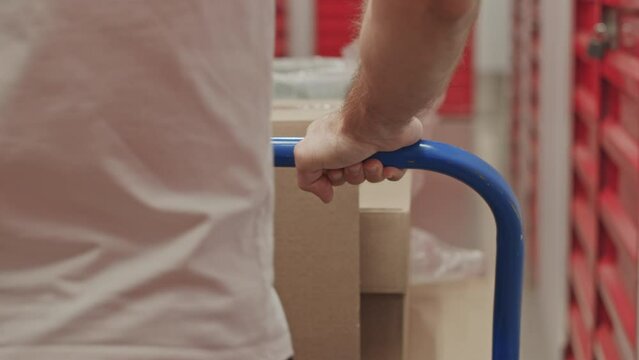 Close up back view shot of unrecognizable man pushing cart loaded with boxes while going to self storage unit in warehouse